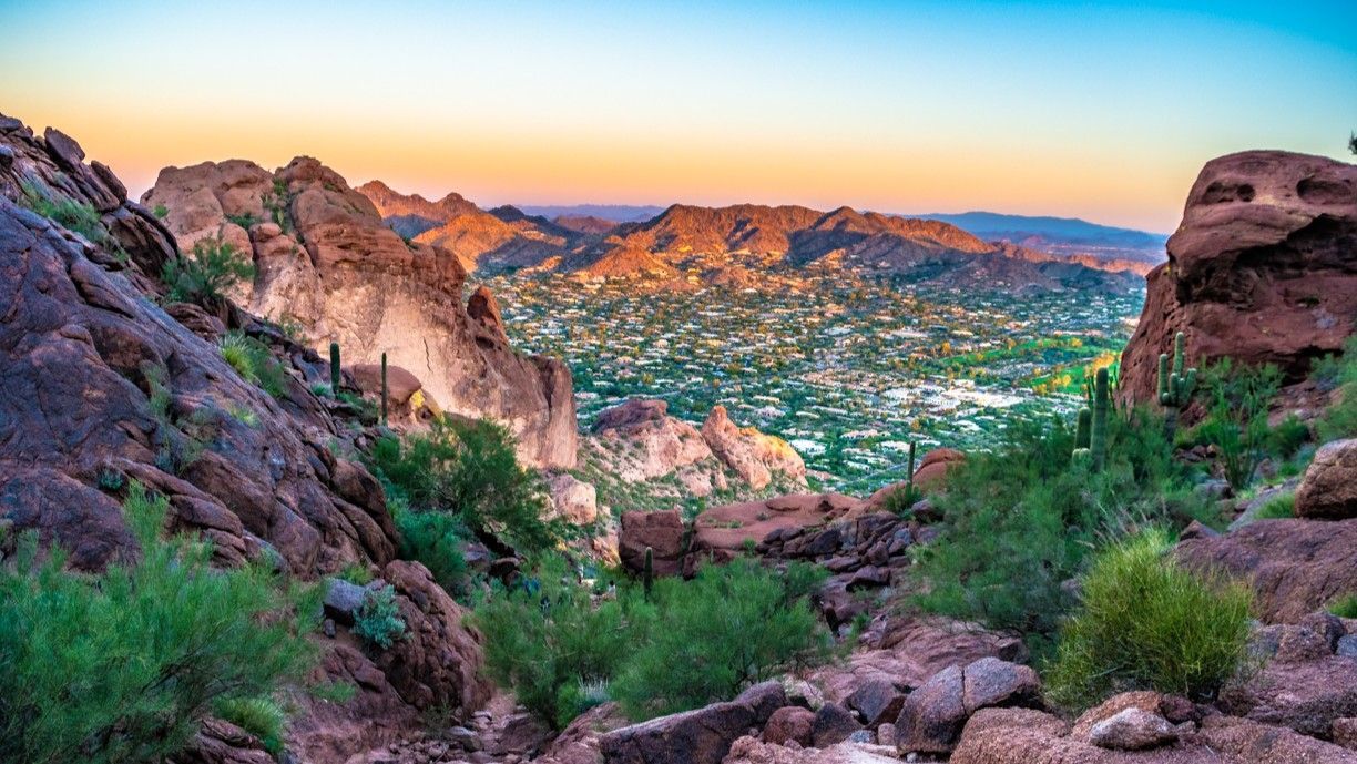 View of a city from a rocky mountain during sunrise, with colorful sky and vegetation.