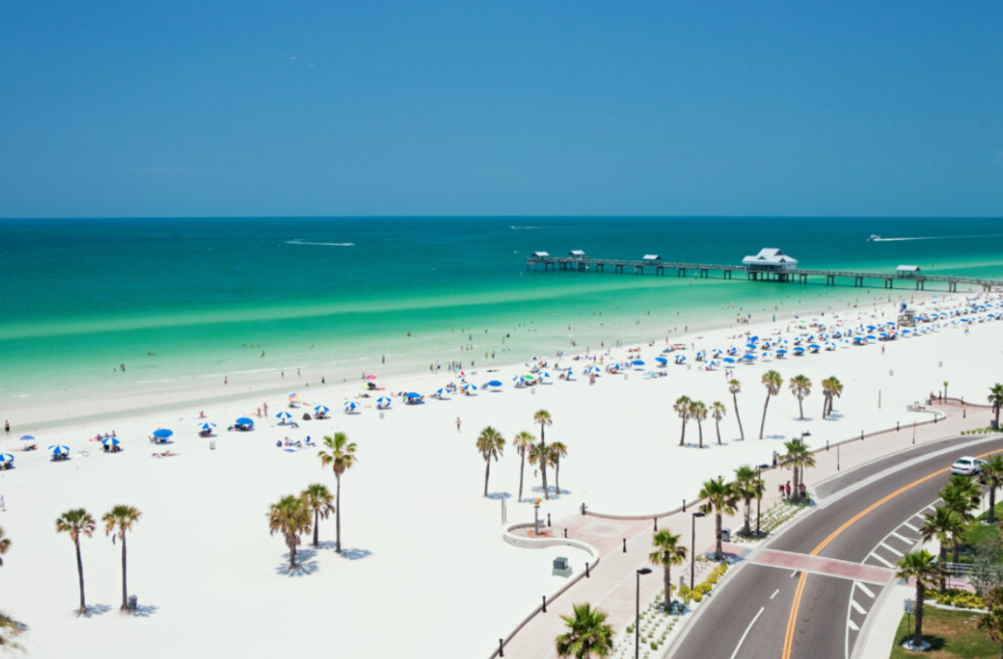 Beach scene with white sand, turquoise water, pier, palm trees, and road.