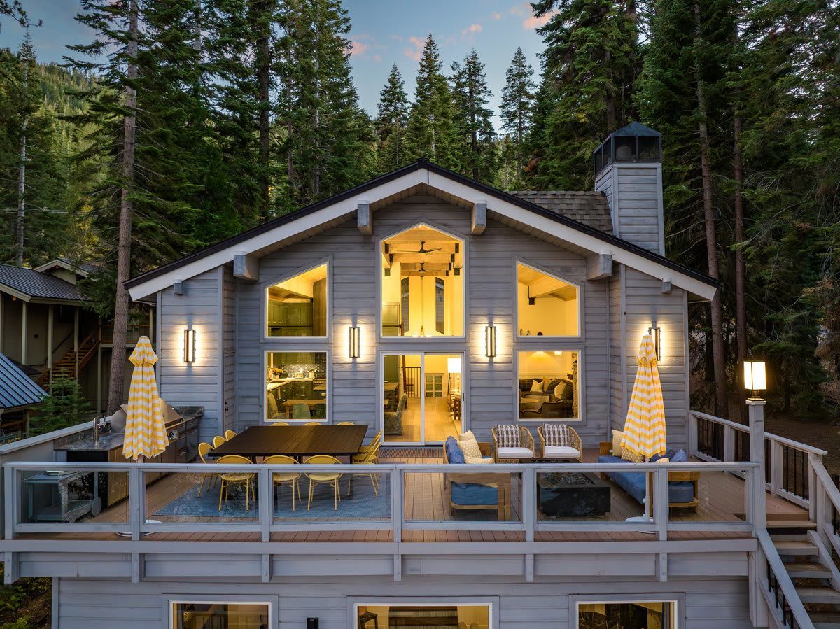 Cozy mountain cabin at dusk with lit windows, deck seating, and tall pines surrounding it