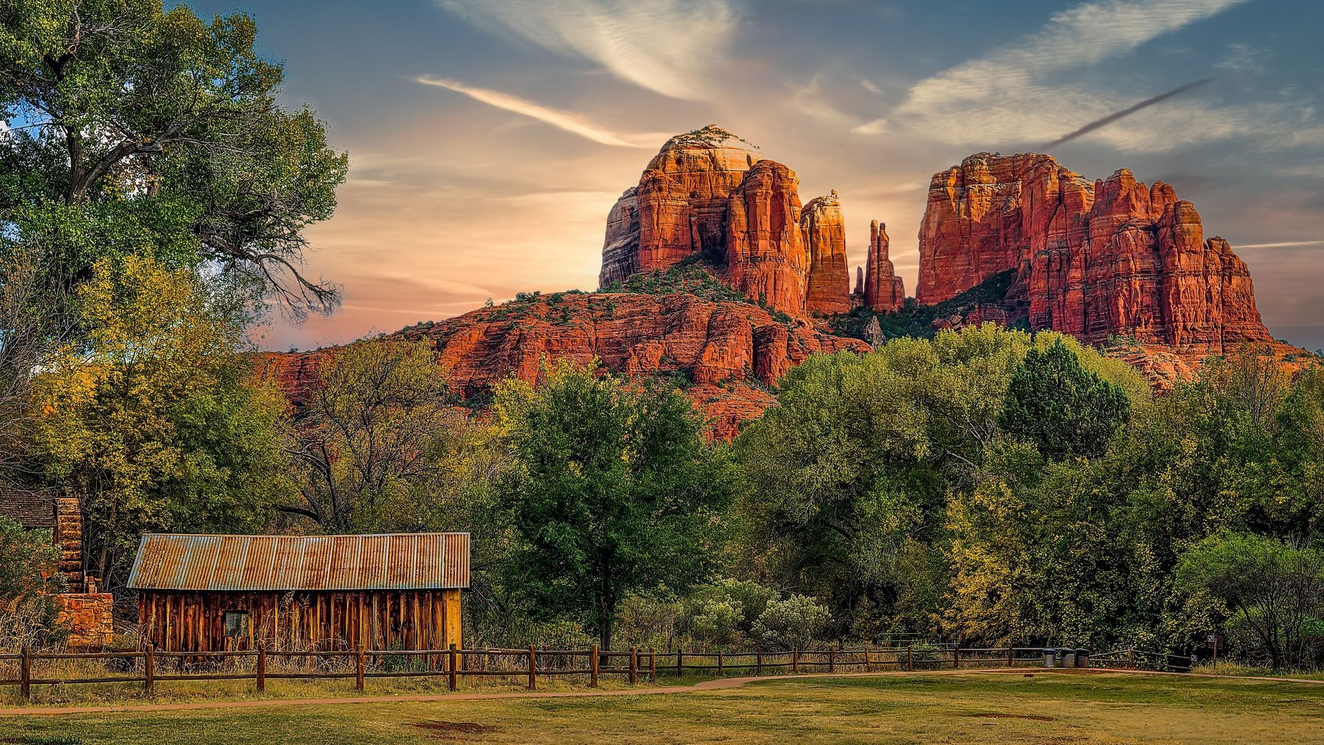 Red rock formations rising behind a wooden structure and trees in a sunny landscape.