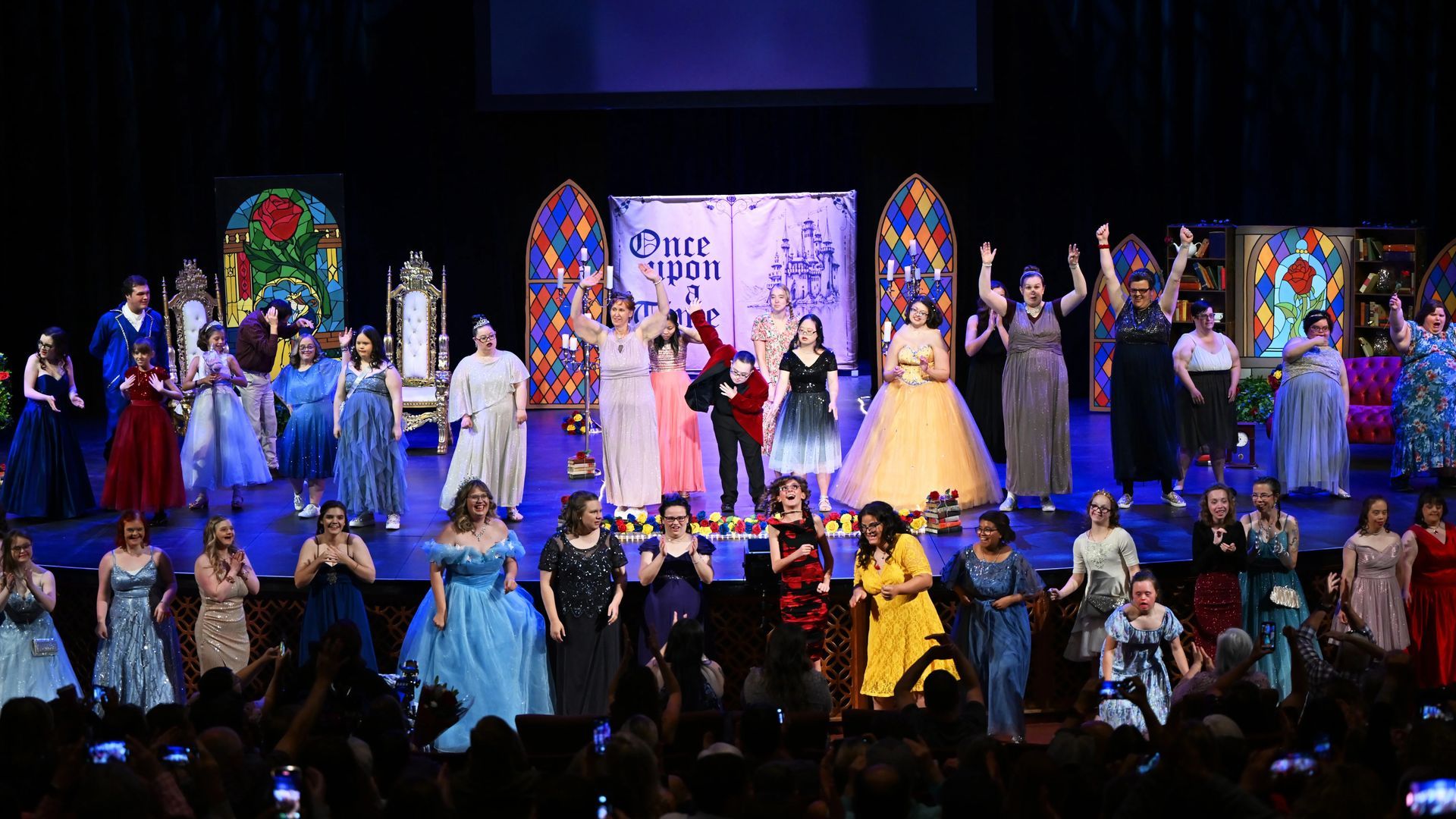 Musical cast takes a bow on stage. Colorful gowns, decorated backdrop, and audience applauding.
