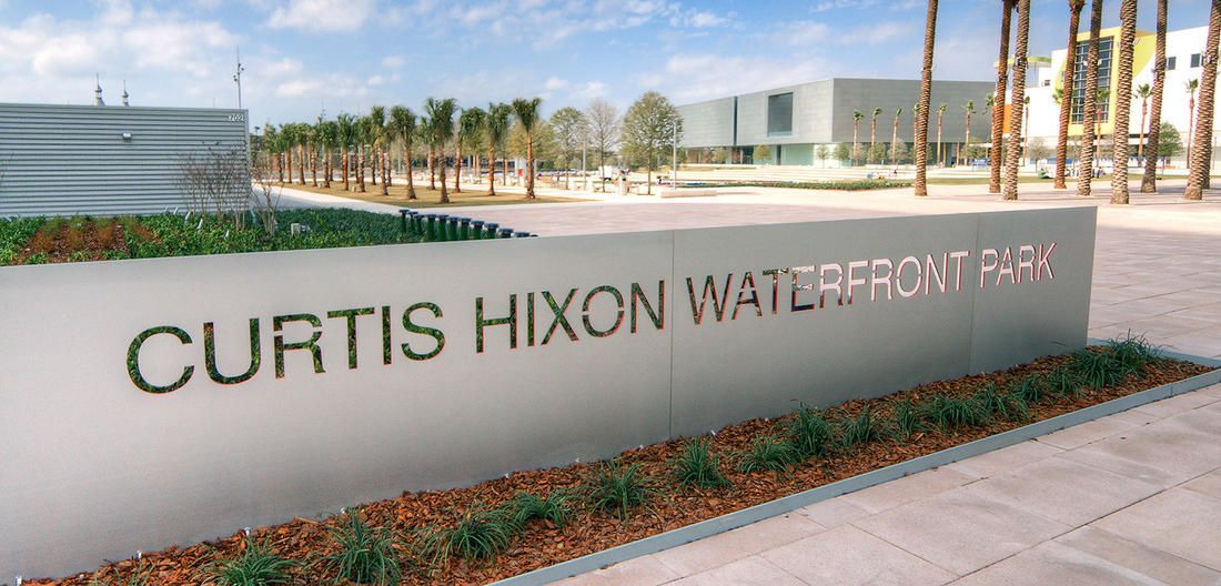 Curtis Hixon Waterfront Park sign. A sign in front of a park with lush greenery and a cityscape in the background.
