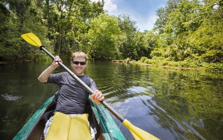 Man kayaking on a river, smiling, holding a yellow paddle. Trees line the riverbank, sunny.