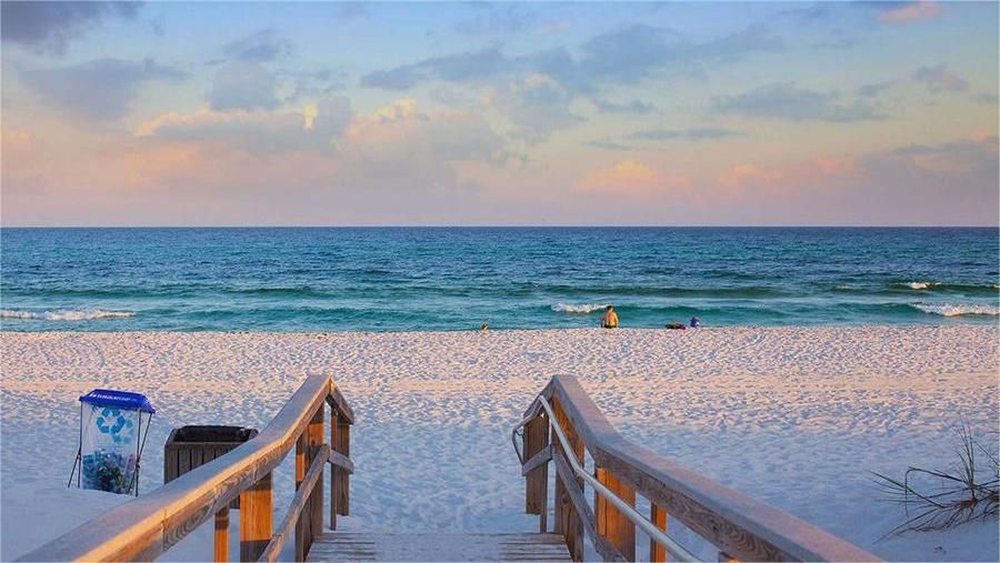 Beach scene at sunset. Wooden walkway leads to white sand and turquoise ocean, with pink and blue sky.