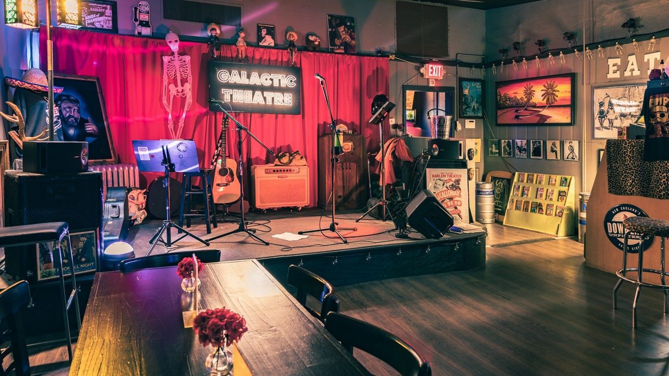 Indoor music venue with stage, instruments, and a performer. The stage has red curtains and the word 