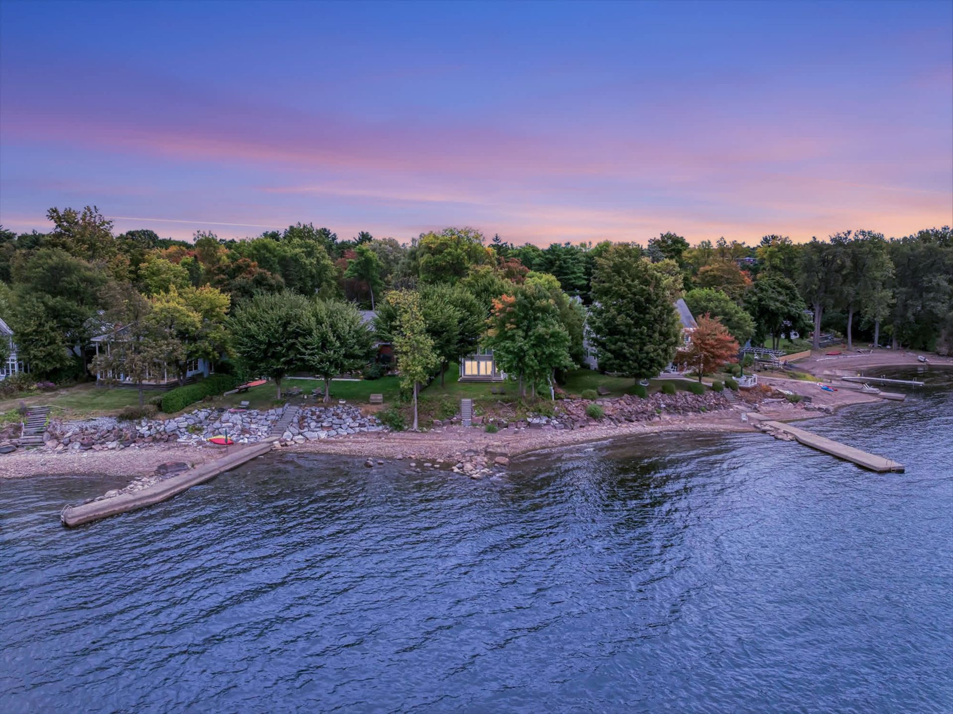Coastal shoreline with dense green trees, rocky docks, and calm blue water at sunset