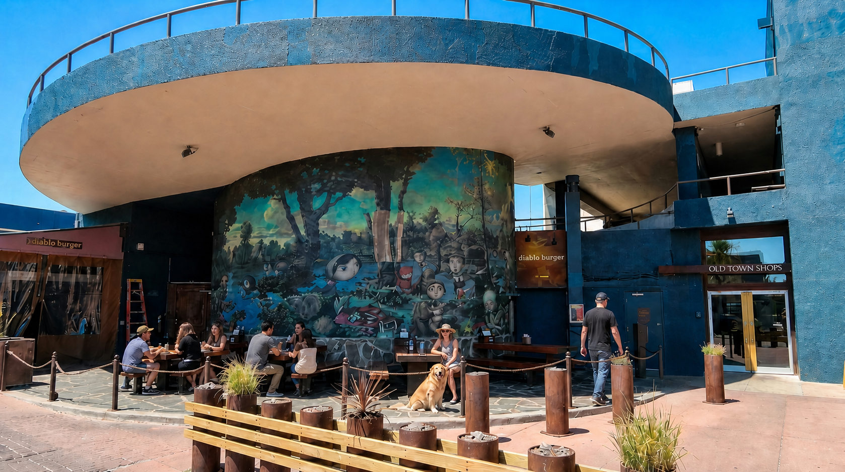People seated outside a colorful building with a large mural and round overhang in bright sunlight