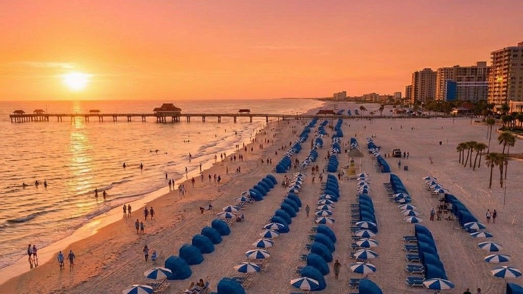 A sunset over a beach, featuring a long fishing pier, sand filled with rows of blue umbrellas, and nearby hotel buildings.