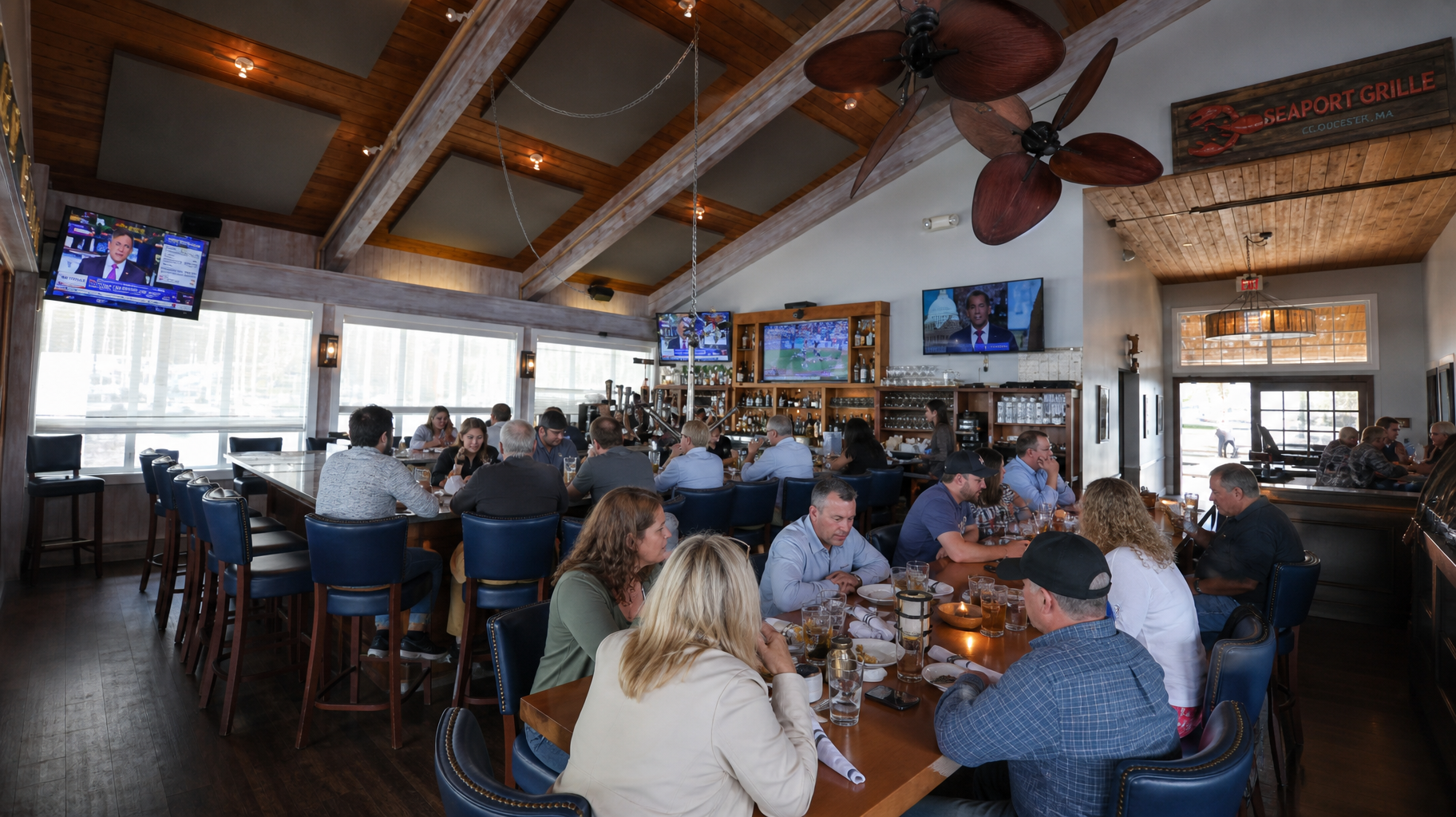 Busy restaurant dining room with patrons seated at tables under ceiling fans and TVs above the bar.