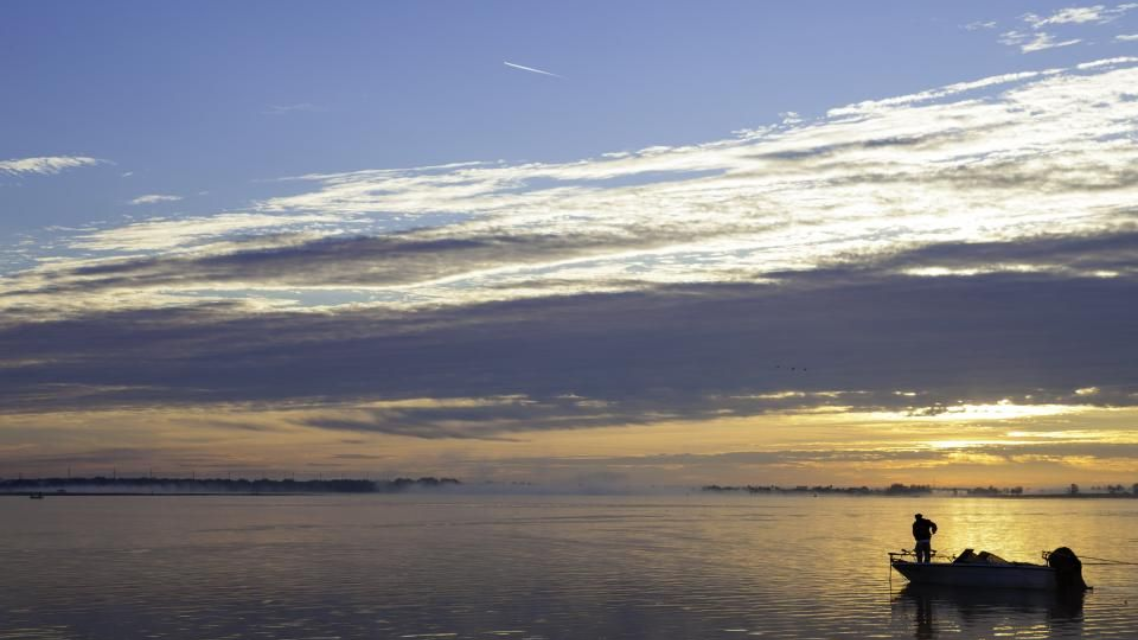 Boat with person fishing on calm water at sunrise; golden light, cloudy sky.