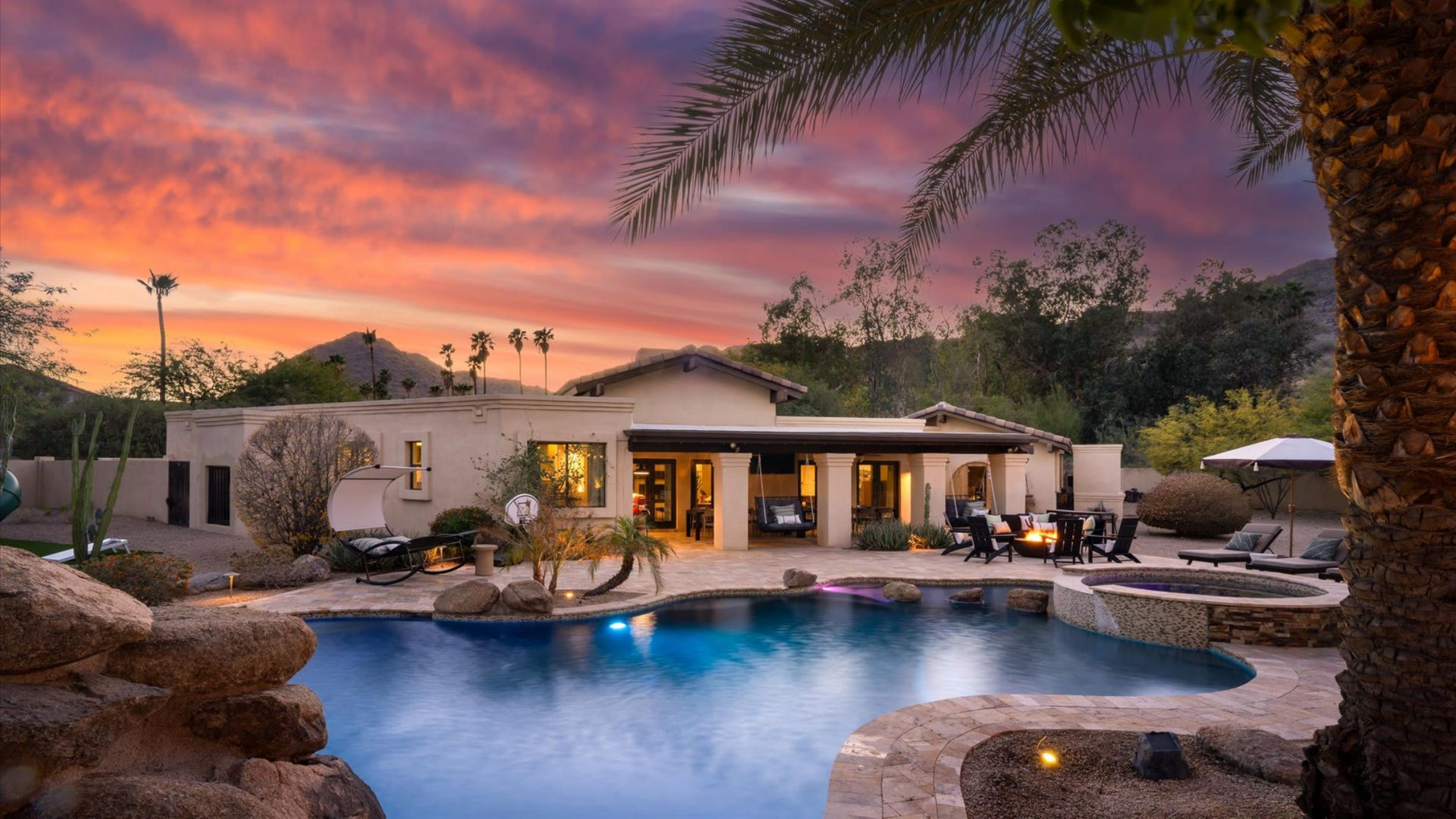 Desert home with lit patio and blue pool at sunset, palm trees silhouetted against a pink-purple sky