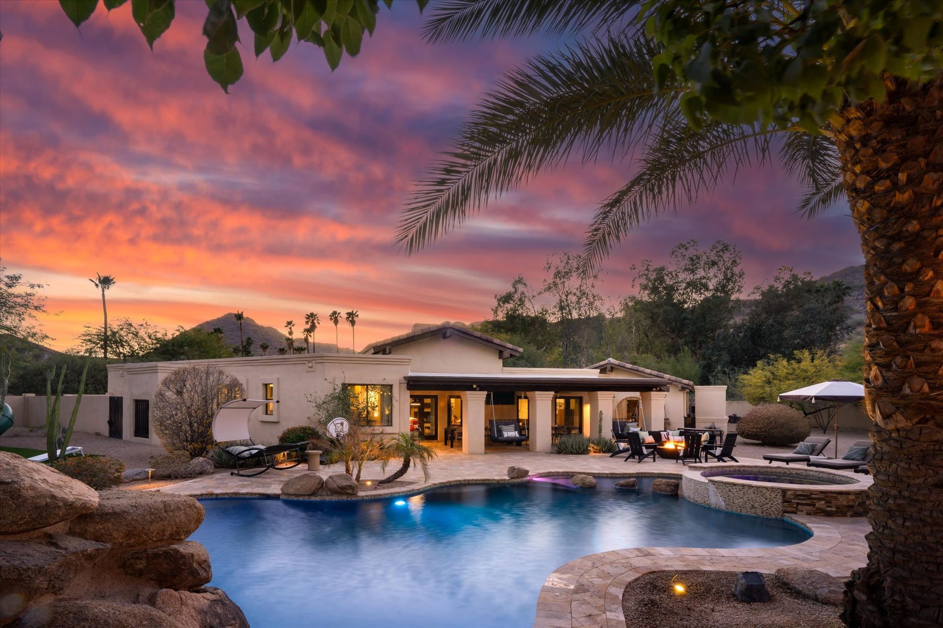 Desert backyard with a blue pool, palm tree, patio seating, and mountain views under a sunny sky