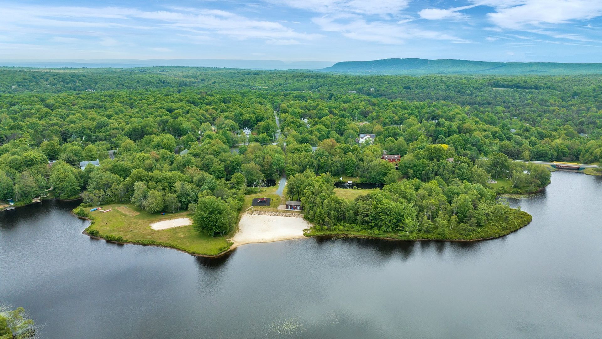 Aerial view of a sandy beach on a lake peninsula, surrounded by green trees, under a blue sky.