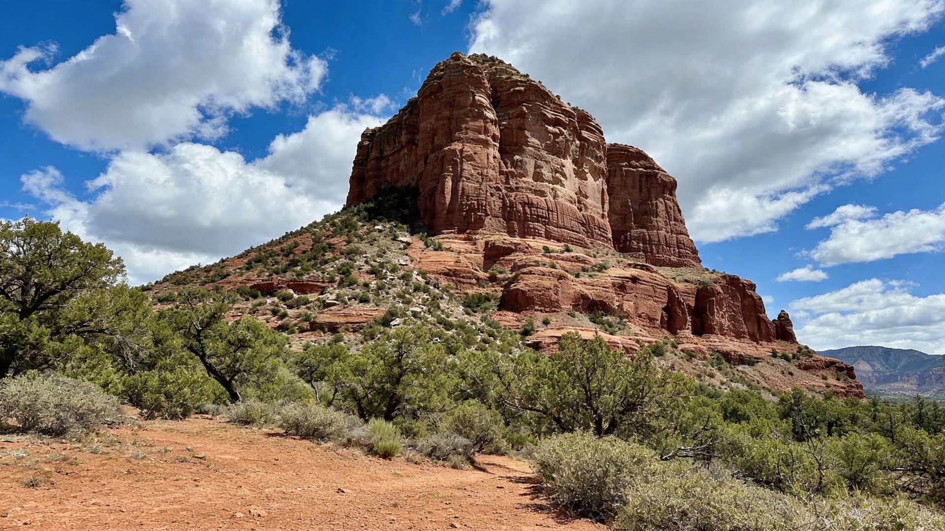 Red sandstone butte under a blue sky with clouds, surrounded by desert shrubs and rocky ground