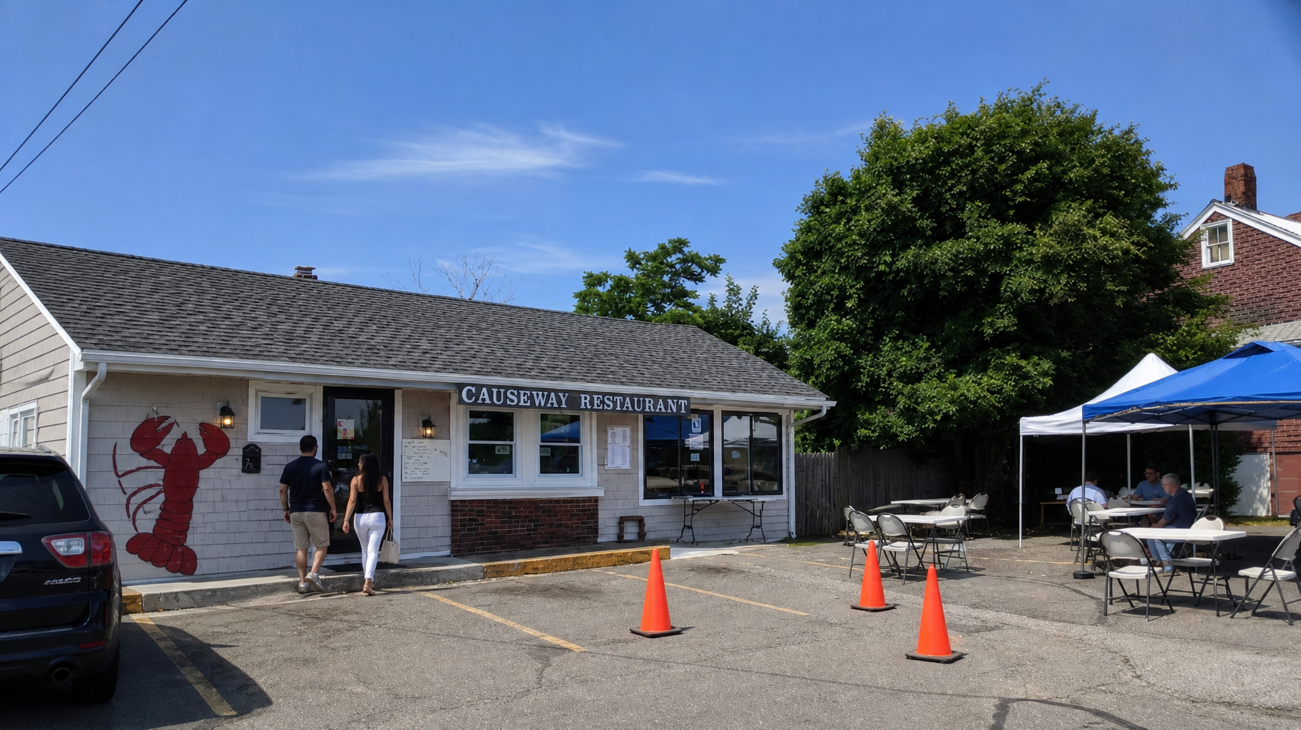 Small roadside cafe with outdoor seating, orange cones, and two people standing by the entrance under a blue sky
