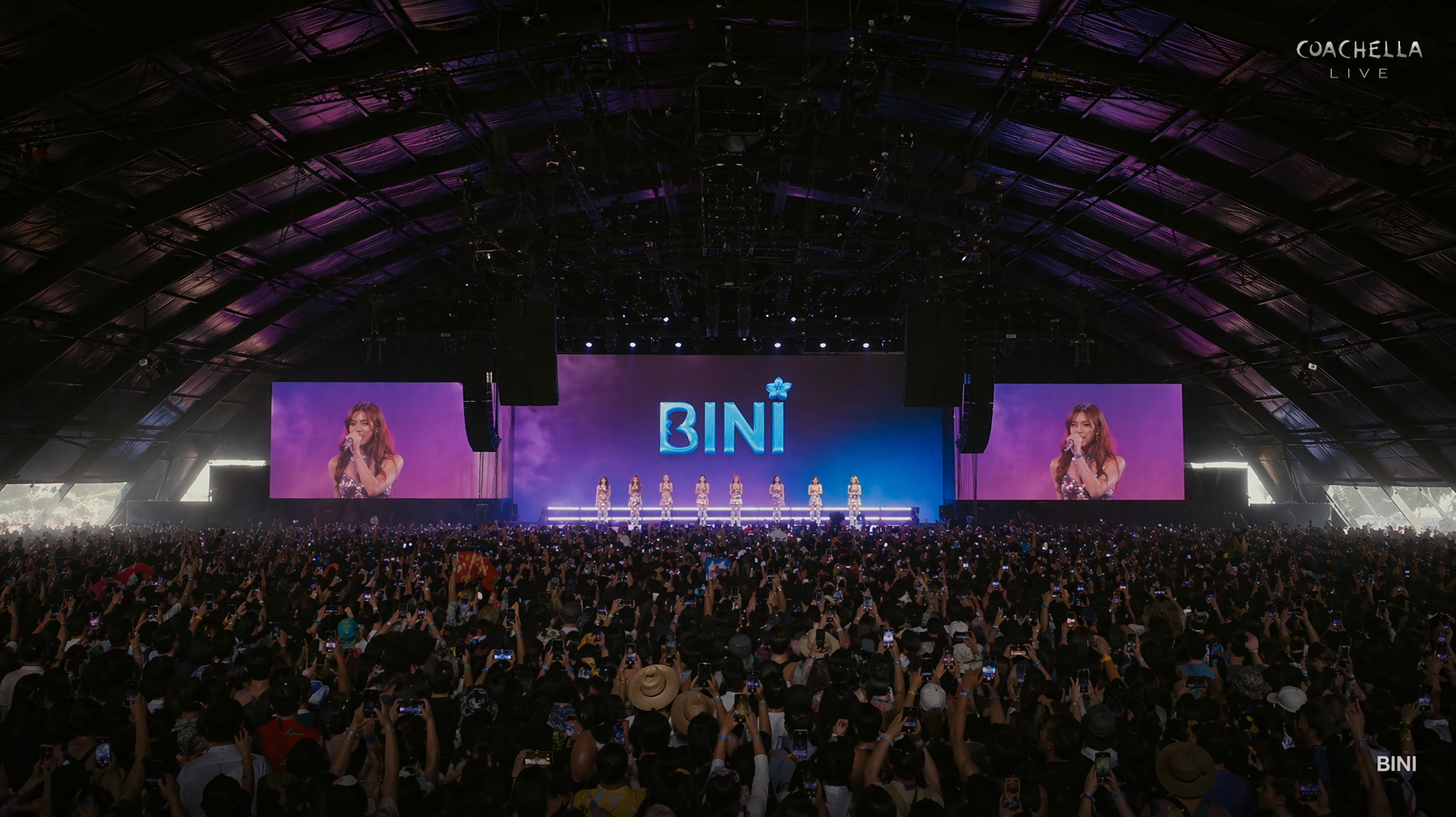 Wide concert stage with “BINI” on a central LED screen and a large cheering crowd under a purple-lit arched roof