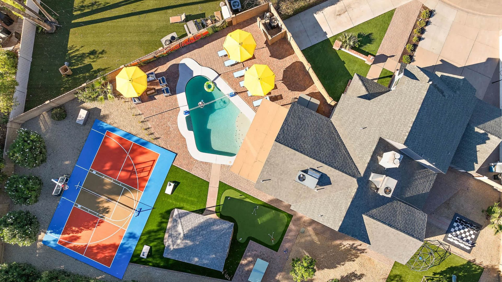 Aerial view of a backyard with a pool, patio seating, hot tub, trampoline, and house roof.