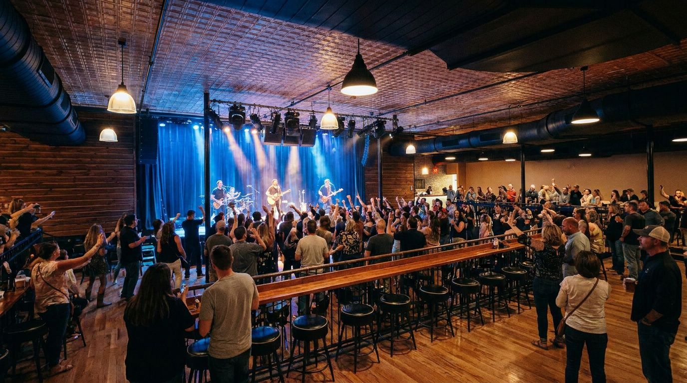 Crowded concert hall with a live band on stage, blue lights, and audience standing at long tables