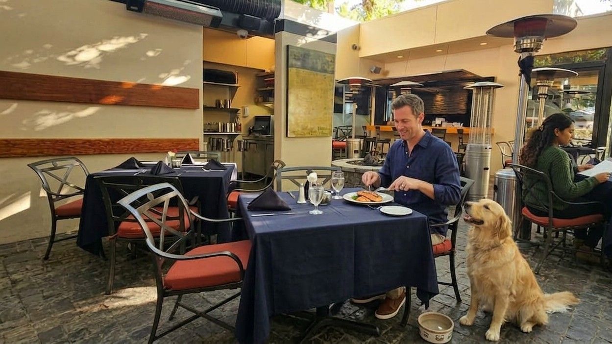A man dines at an outdoor restaurant table with a golden retriever sitting beside him, while a woman sits at a table nearby.
