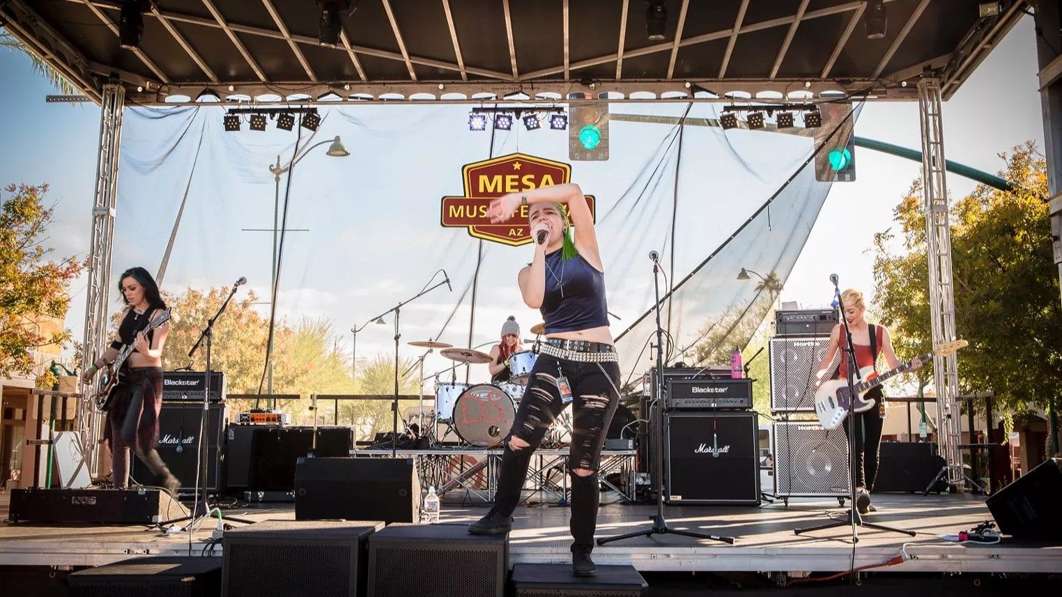 A rock band performs on an outdoor stage: singer with green hair, two guitarists, drummer.