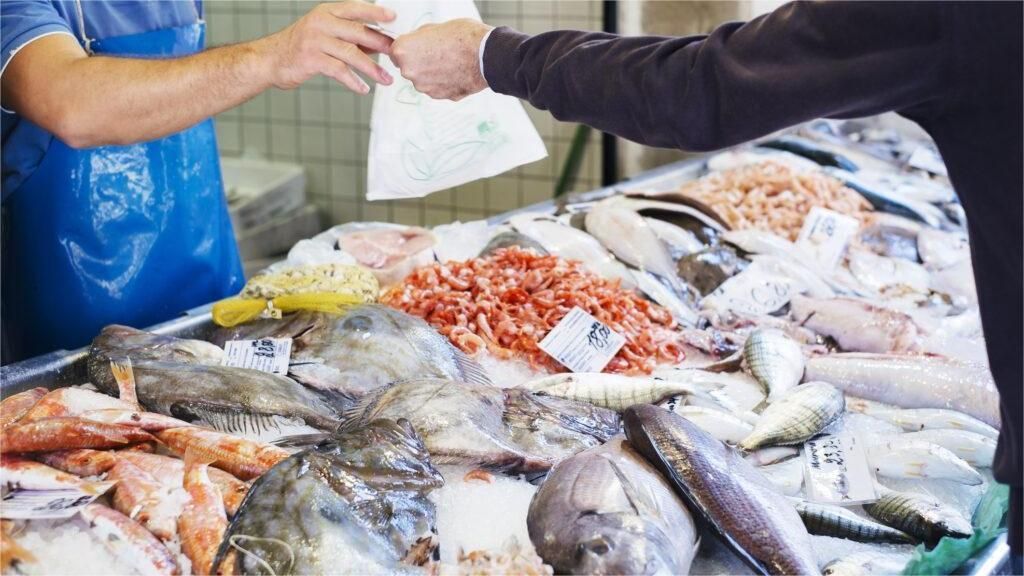 Person buying seafood at a fish market; hands exchanging a bag. Various fish displayed on ice.