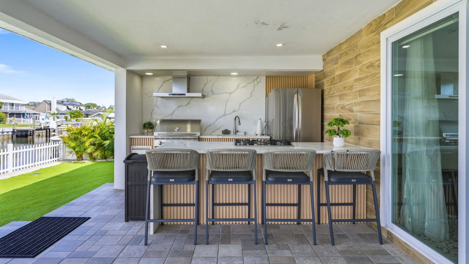 Outdoor kitchen with a bar and stools, next to a waterfront lawn.
