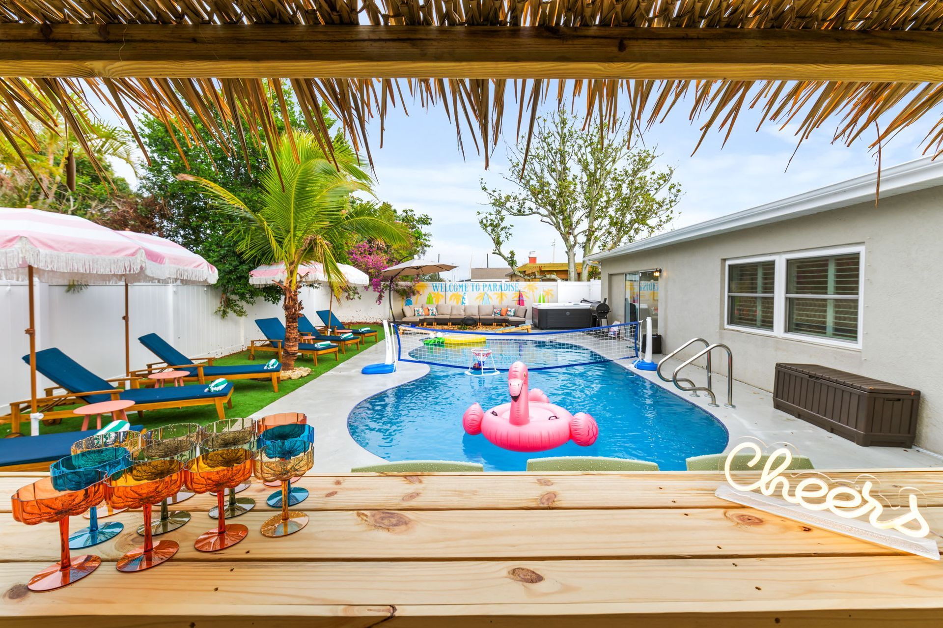 Backyard pool with pink floats, lounge chairs, and a patio bar under a thatched roof