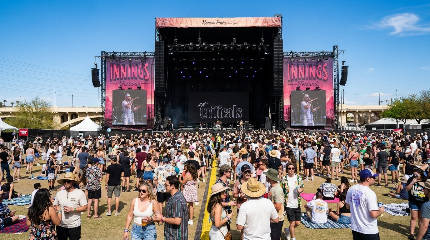 Crowd gathered before a large outdoor concert stage with screens and speakers under a clear blue sky