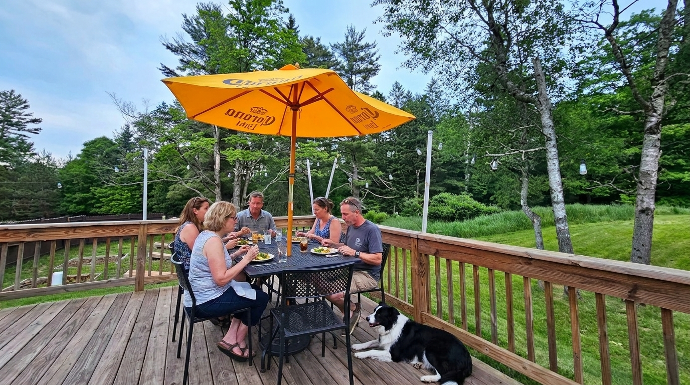 Group sitting around an outdoor table on a wooden deck under an orange umbrella, with a dog nearby and trees around them