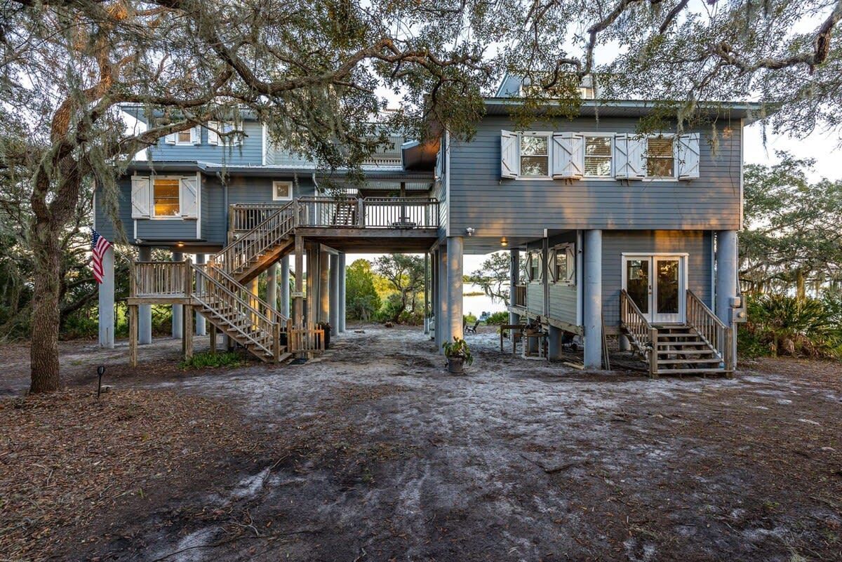 Gray elevated beach house with stairs, covered deck, and large oak tree in front yard