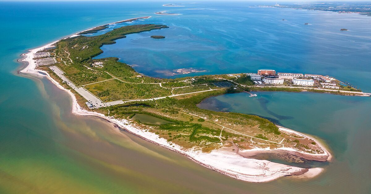 Aerial view of a thin, green island with white beaches surrounded by blue water. Buildings are visible on the right.