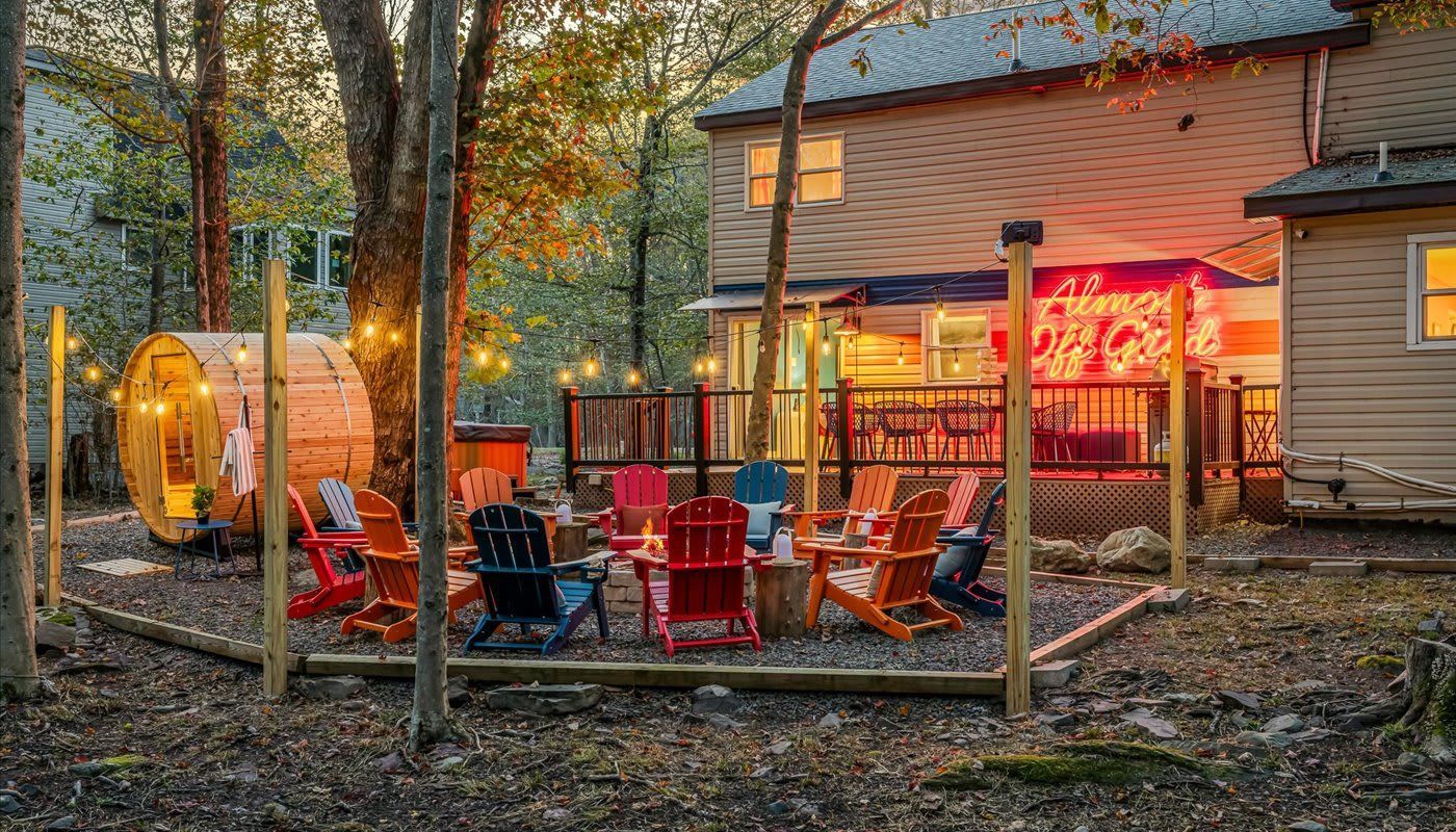 Outdoor gathering area with Adirondack chairs around a fire pit, next to a barrel sauna and a house.