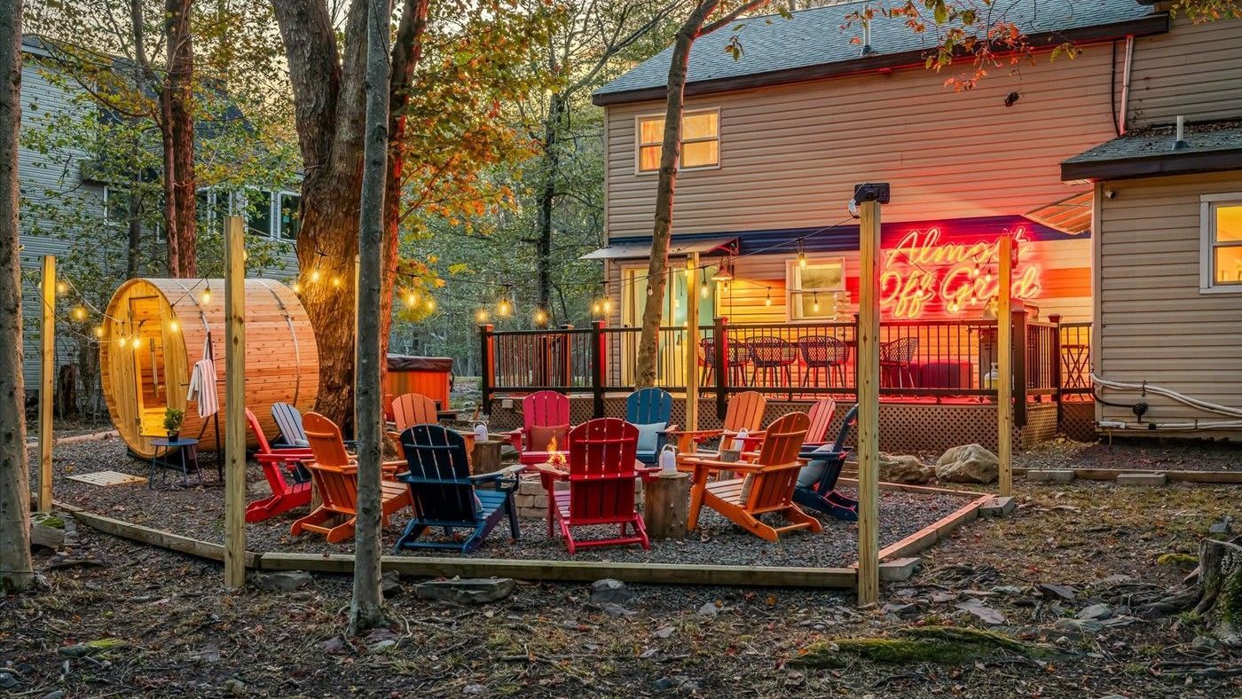 An outdoor fire pit area at dusk with colorful Adirondack chairs, string lights, and a nearby barrel-shaped sauna.