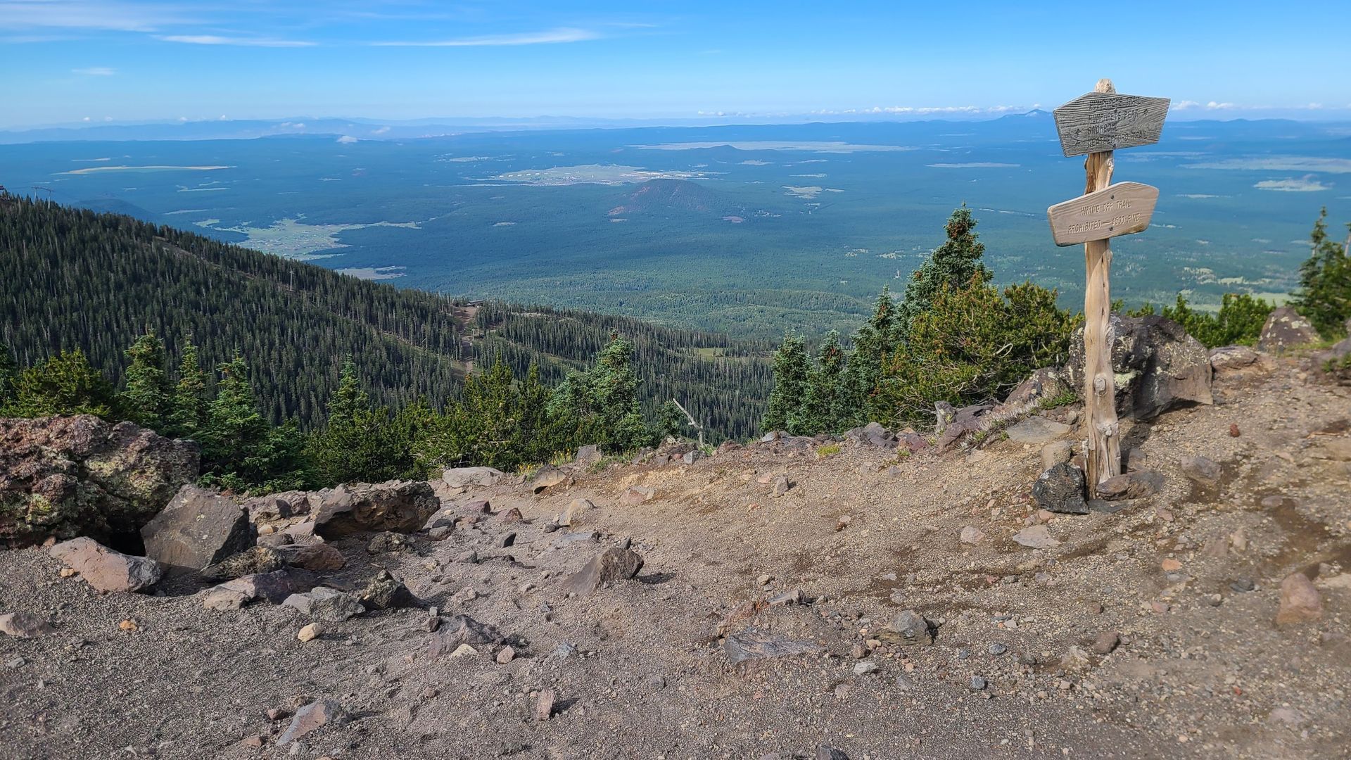 View from mountain peak with a wooden signpost. Distant trees and town visible under a blue sky.