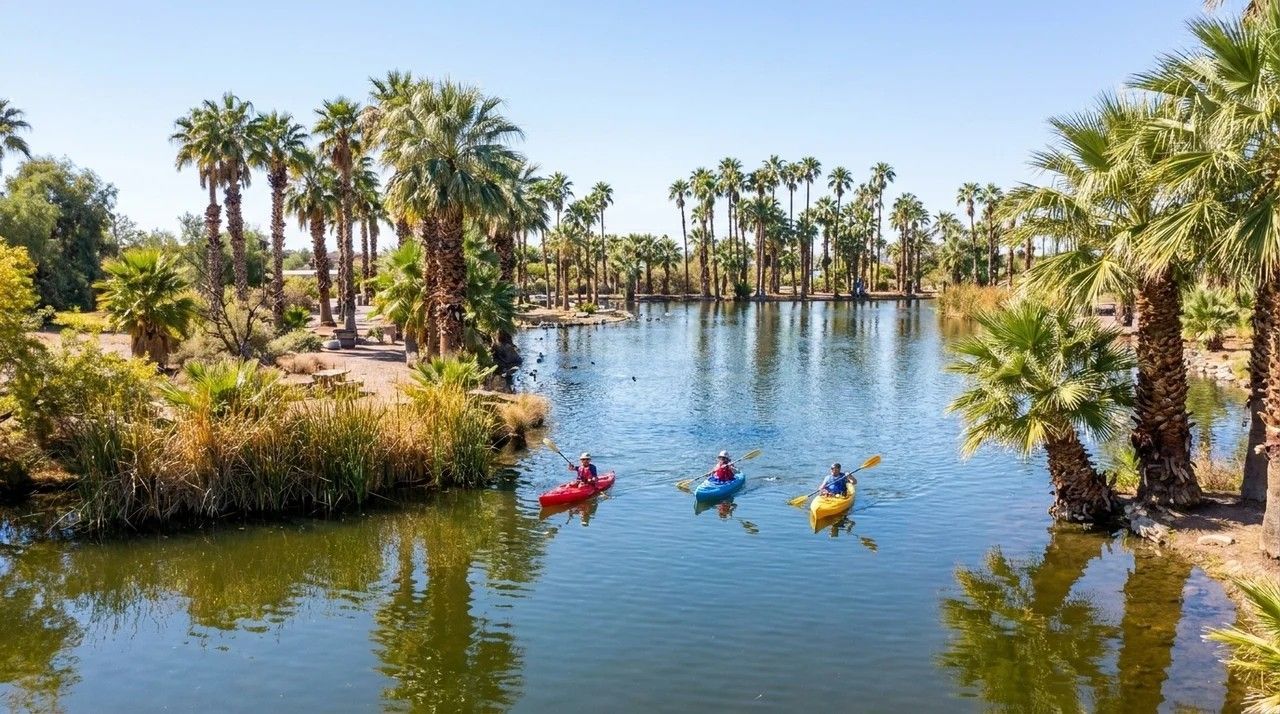 Kayakers paddling on a calm desert oasis lake lined with palm trees under a blue sky