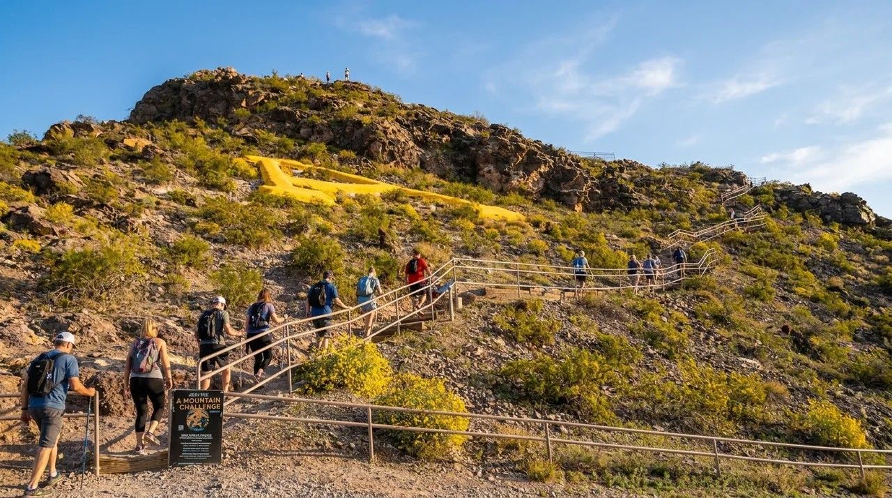 Hikers ascending a rocky hillside trail with wooden railings under a bright sky