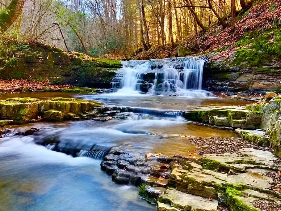 Waterfall cascading over layered rocks into a stream in a wooded area; sunlight filters through trees.