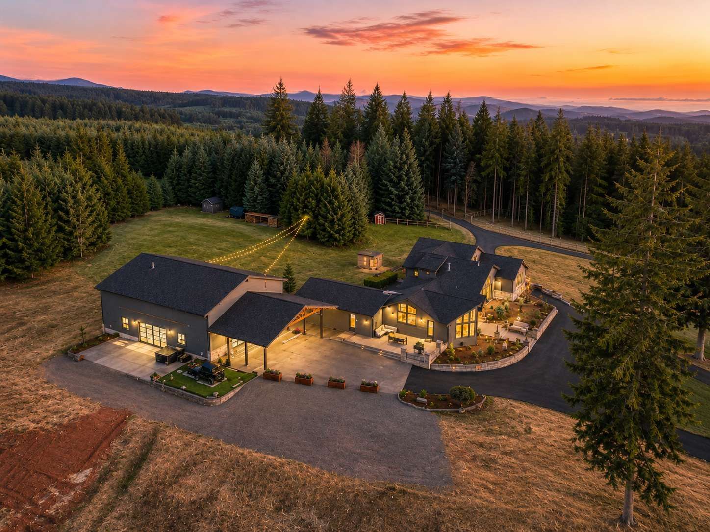 Modern home with warm lights at sunset, surrounded by pine trees and open fields.