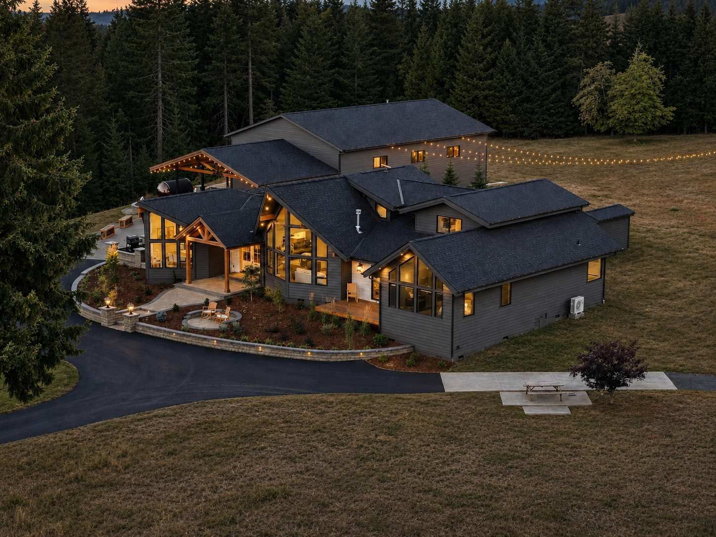 Large modern house with warm lights at dusk, surrounded by trees and a curved driveway