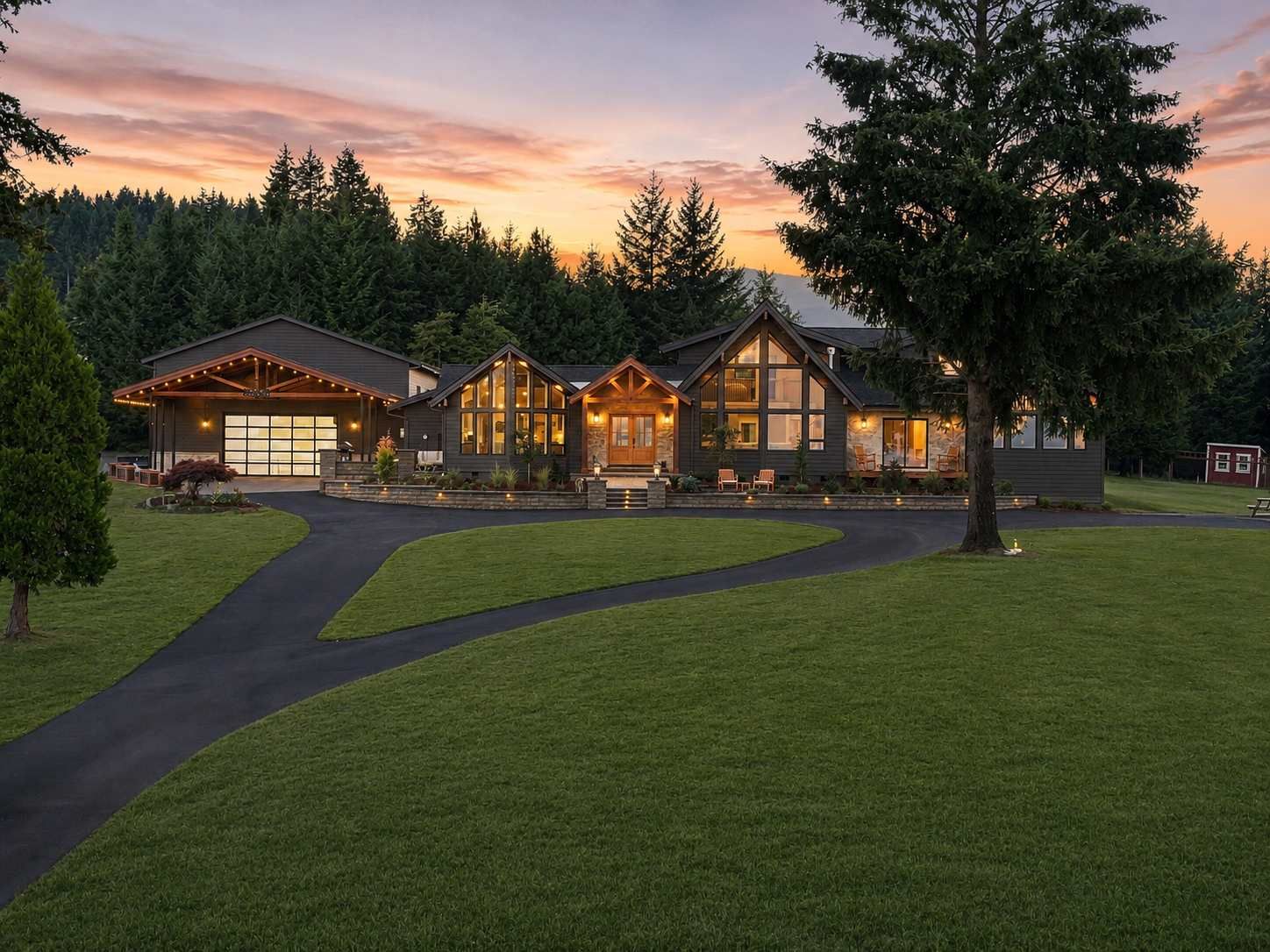 Large illuminated house at dusk with a wide green lawn and driveway under a pink sunset sky