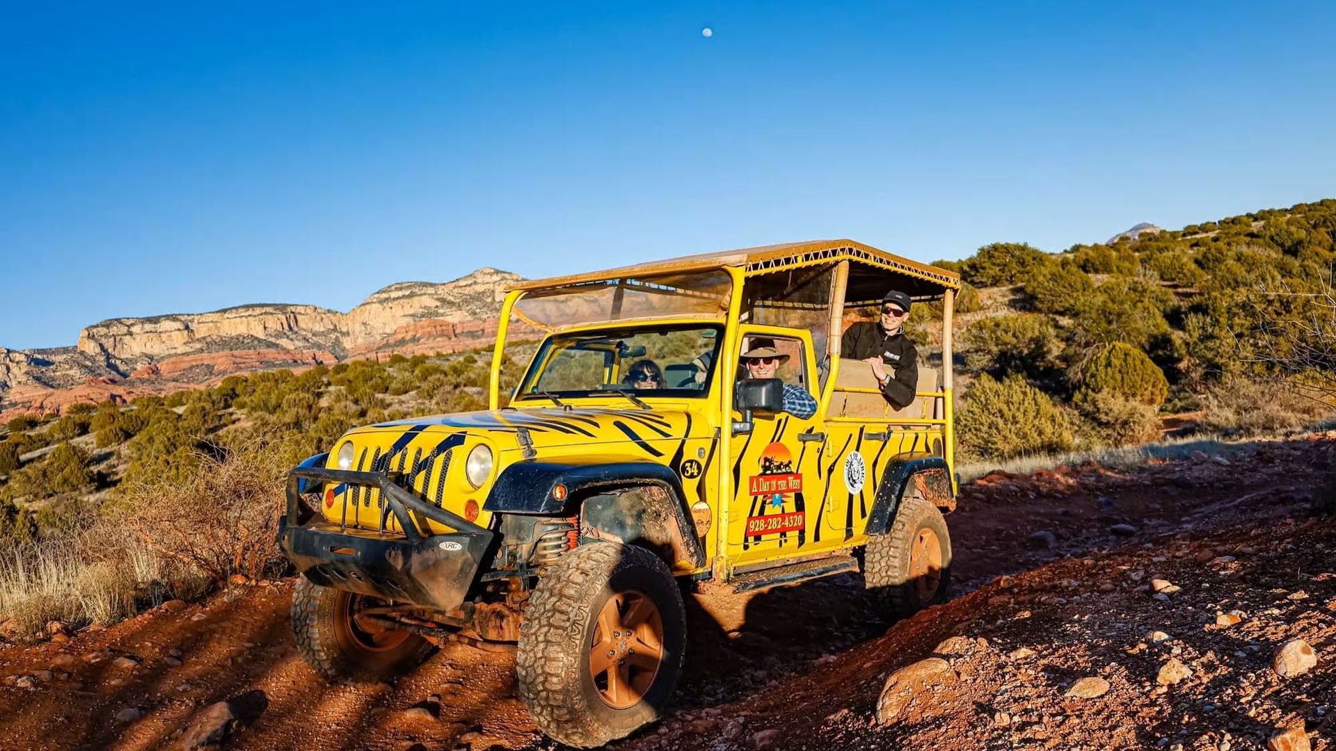 Yellow Jeep on a dirt road, with passengers, surrounded by red rock formations and vegetation.