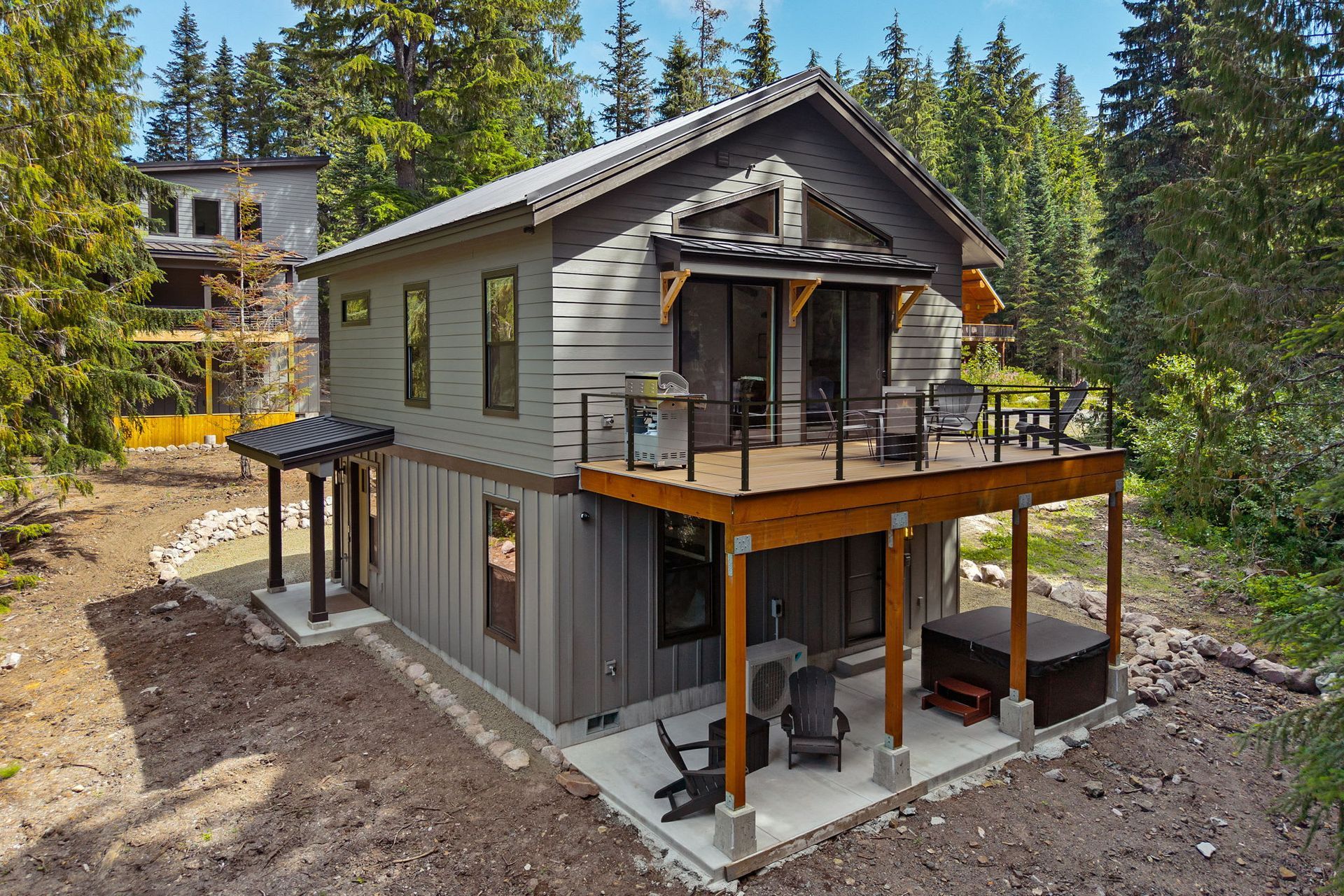 Modern two-story cabin with covered porch and deck in a forested, partially built setting