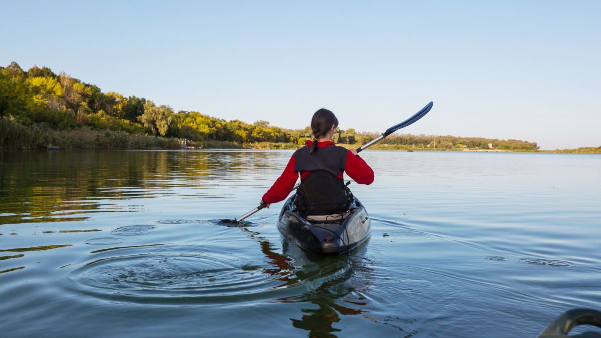 Person kayaking on calm water towards a treeline under a blue sky.