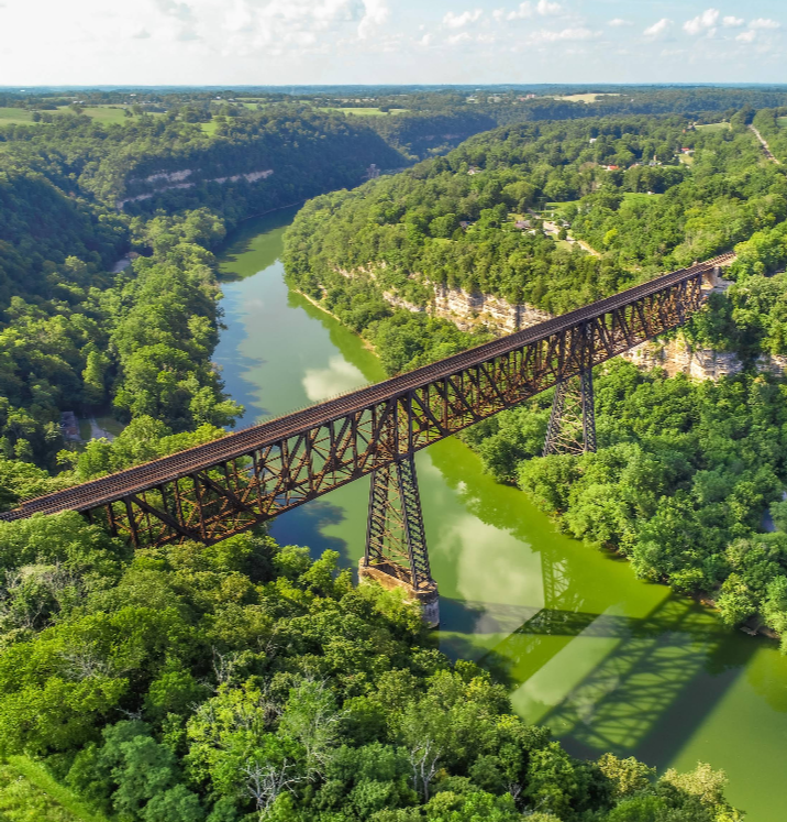 Railroad bridge spanning a green river through a lush, forested valley.