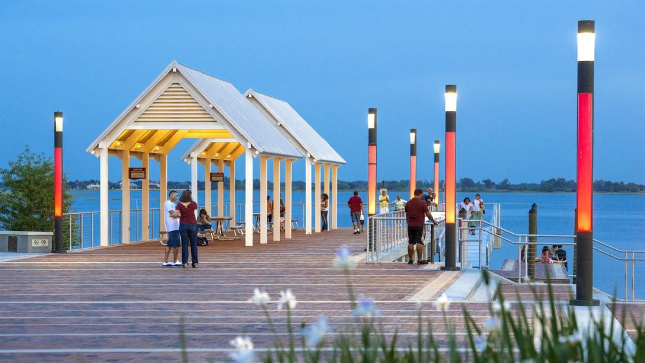Pier with a white pavilion and colorful lights, people walking and enjoying the waterfront at dusk.