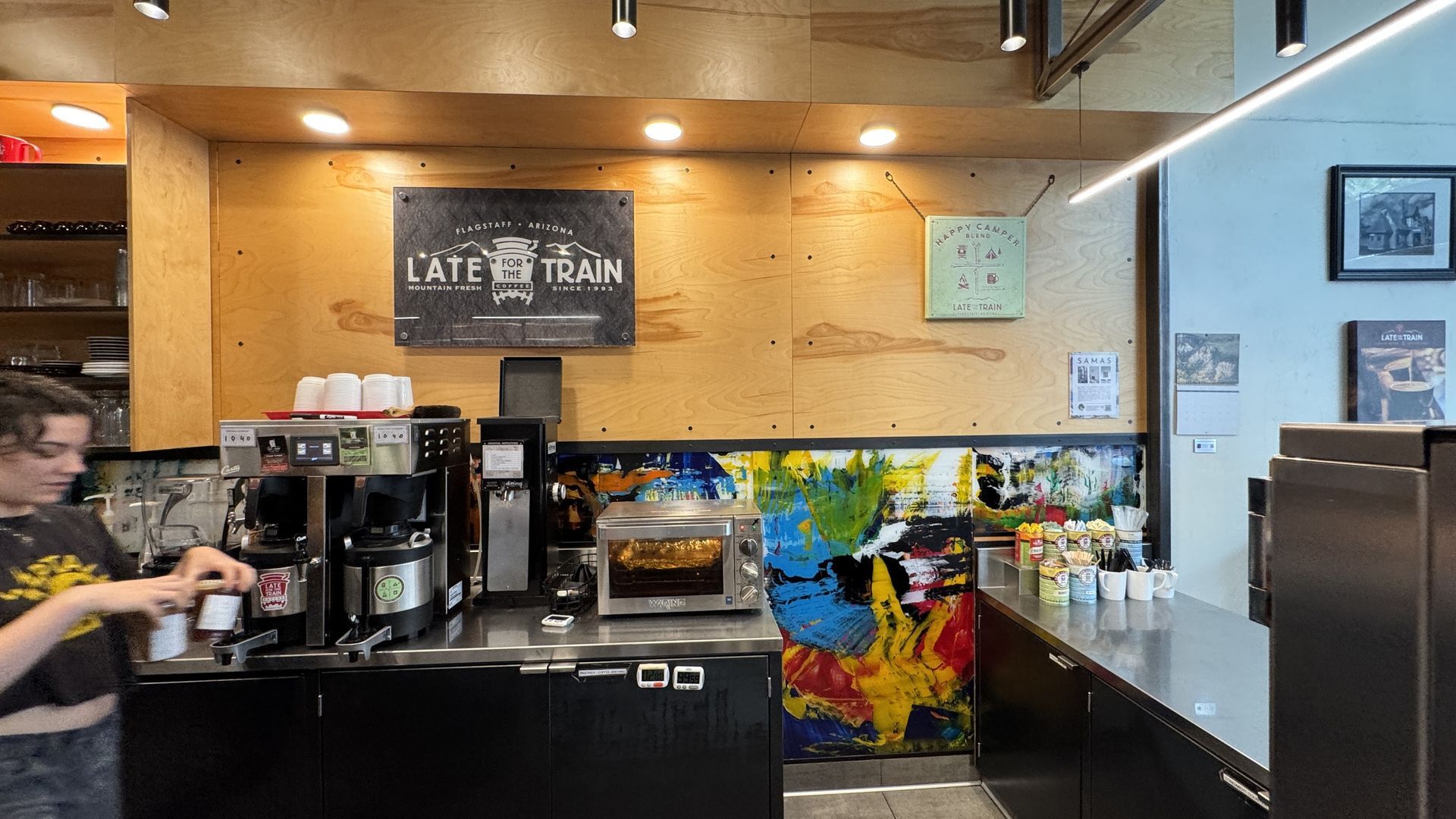 A person prepares a drink behind the counter of a café with plywood walls, a colorful art backsplash, and a menu sign.