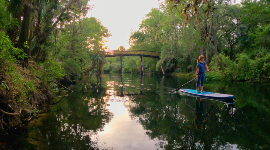 Person on paddleboard, paddling down a calm river under a wooden bridge at dusk, surrounded by trees.