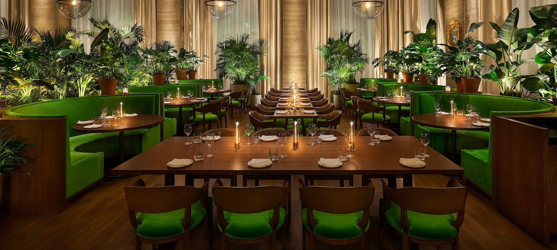 Interior of a restaurant with green booths, wooden tables, and plants. Warm lighting and beige curtains in the background.