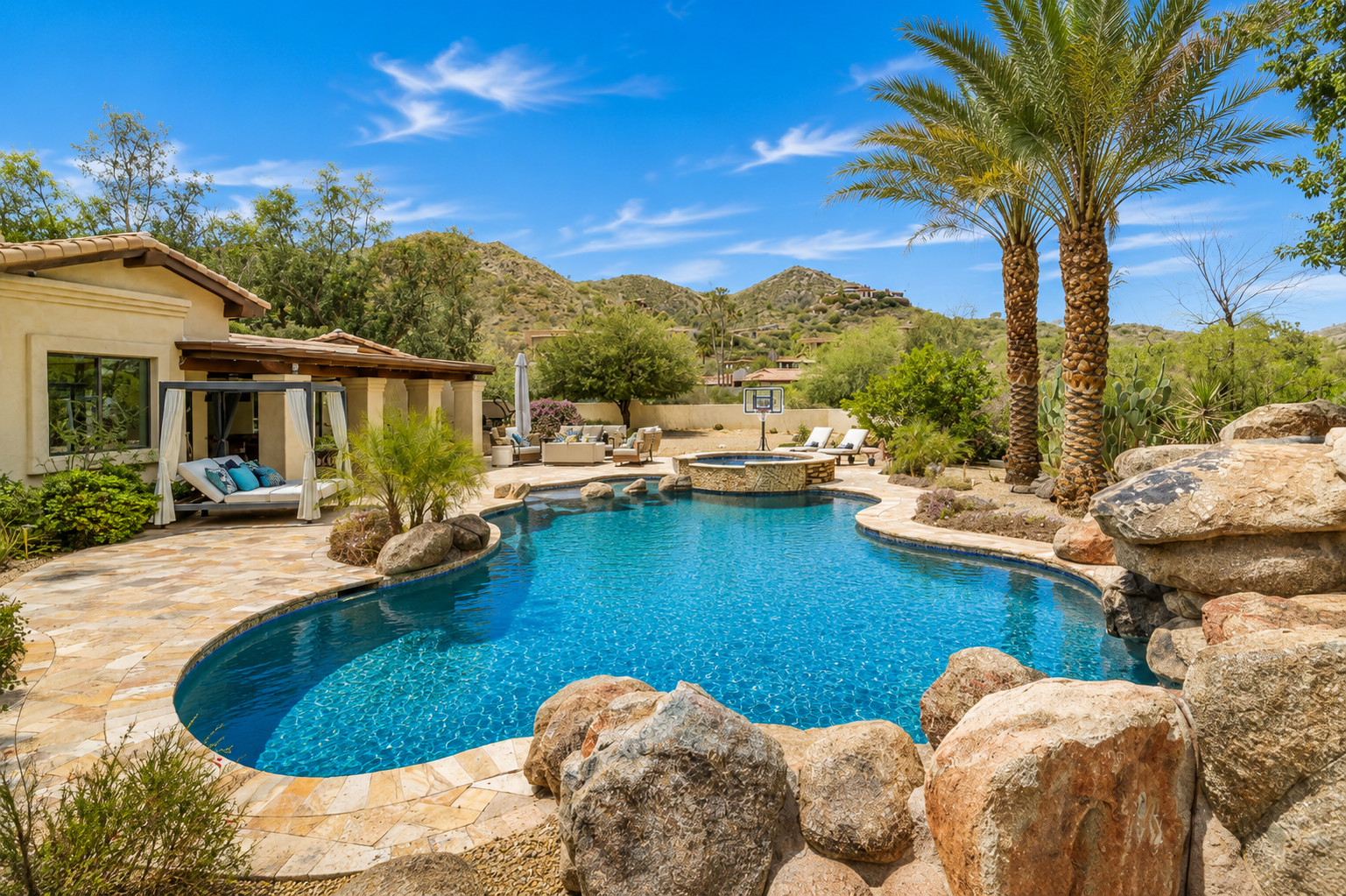 Desert backyard with a turquoise pool, rock landscaping, palm trees, and a patio house under a blue sky