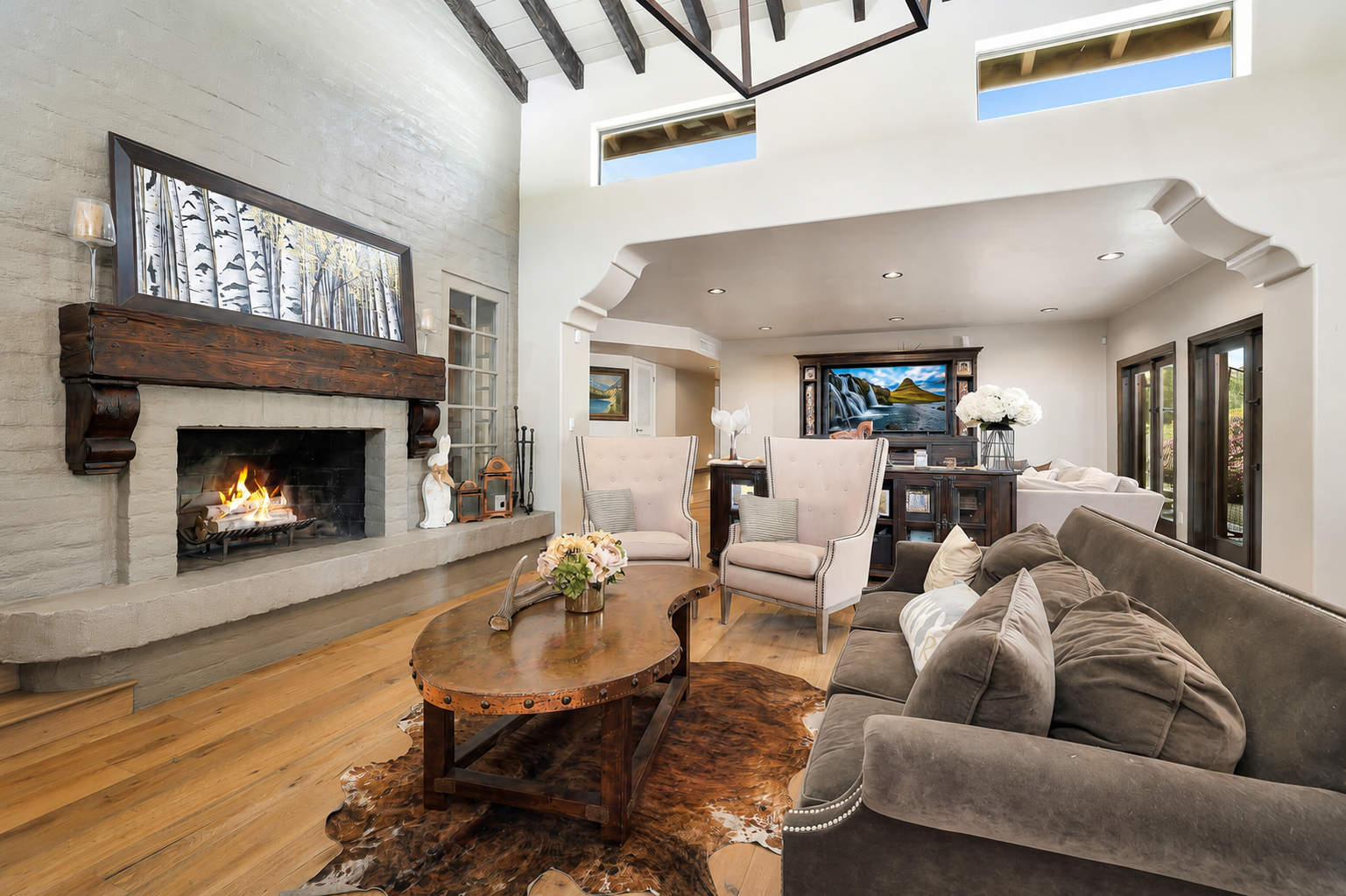 Bright living room with stone fireplace, gray sofa, wooden coffee table, and large windows, viewed toward dining area